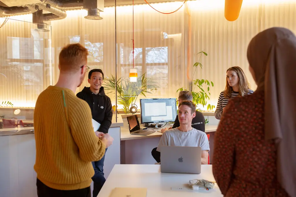 A group of people sitting and standing and chatting around a table, with Apple Mac computers. They are discussing information security, and scaling businesses.