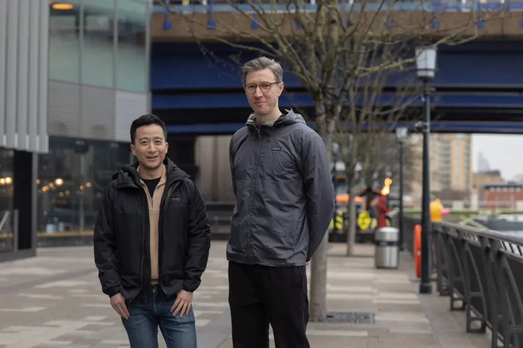 Two people standing outdoors in a city setting, both wearing jackets, with buildings, railings, and a tree-lined walkway in the background. The are reminiscing about the early days of LeftBrain.