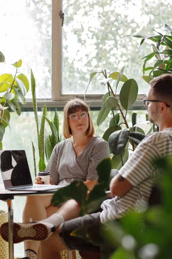 Two people in a Secure Innovation review at a desk, surrounded by large leafy plants, with one holding a takeaway coffee cup and a laptop in view.