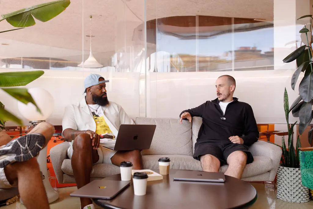 Two people sit on a sofa in a modern office lounge, engaged in conversation. One has a laptop open on their lap, while the table in front holds two more laptops and takeaway coffee cups. Large green plants and stylish lighting fixtures decorate the space.