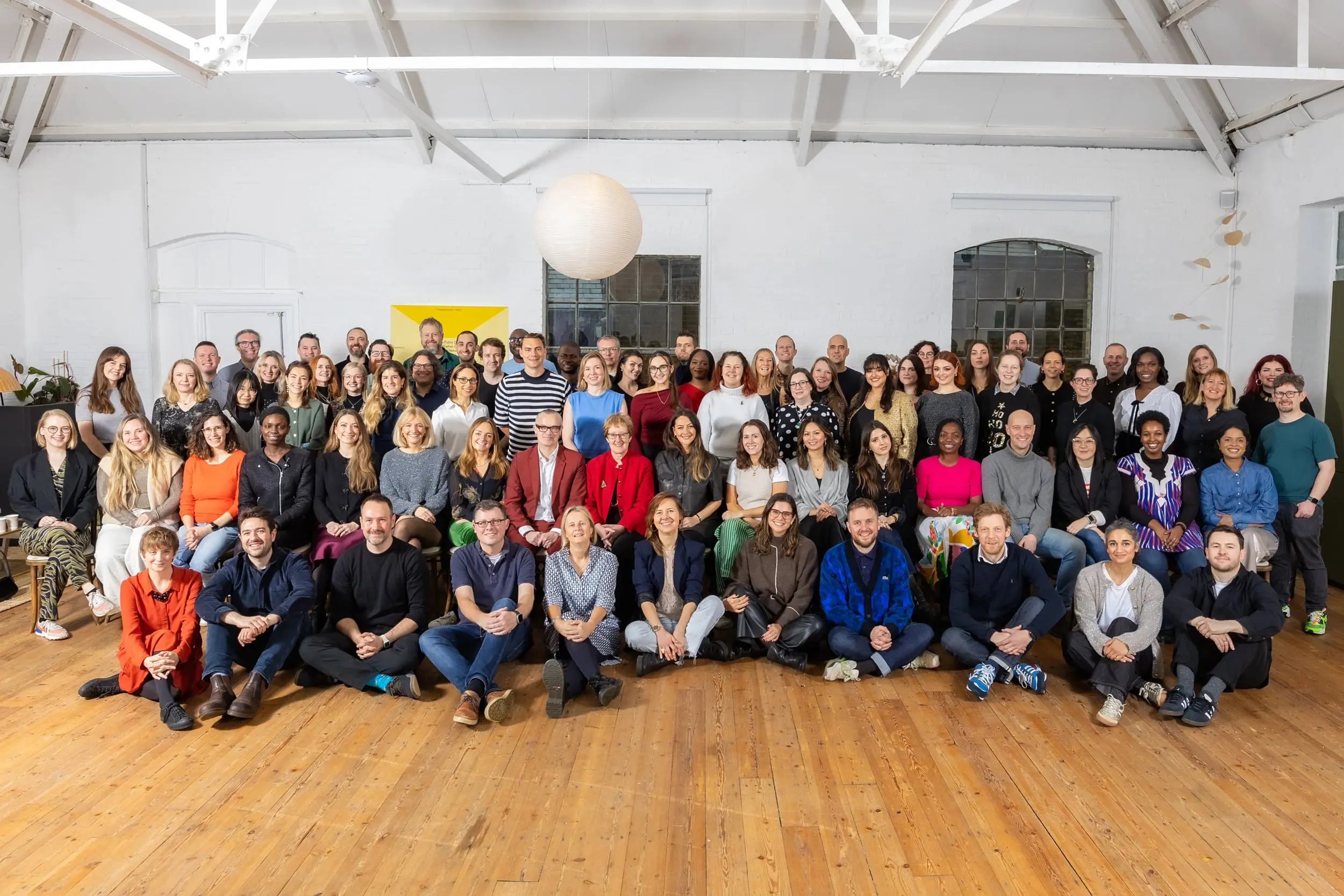 A large group of people gathered for a team photo in a bright, high-ceilinged room with wooden floors and white brick walls. Everyone is smiling and seated or standing in rows, with a round paper light hanging above and colourful artwork in the background.