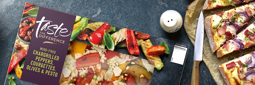 The image shows a boxed supermarket pizza from the "Taste the Difference" range. The flavour is “Wood-Fired Chargrilled Peppers, Courgettes, Olives & Pesto” on a hand-stretched sourdough base. The pizza box is placed on a dark stone surface alongside a sliced cooked pizza topped with red onion and bacon, a knife, and a set of salt and pepper shakers.