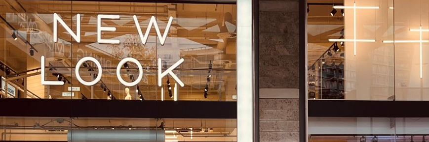 Storefront with large glass windows featuring the bold text “NEW LOOK” in white capital letters; the interior shows modern lighting, mannequins, and retail displays.