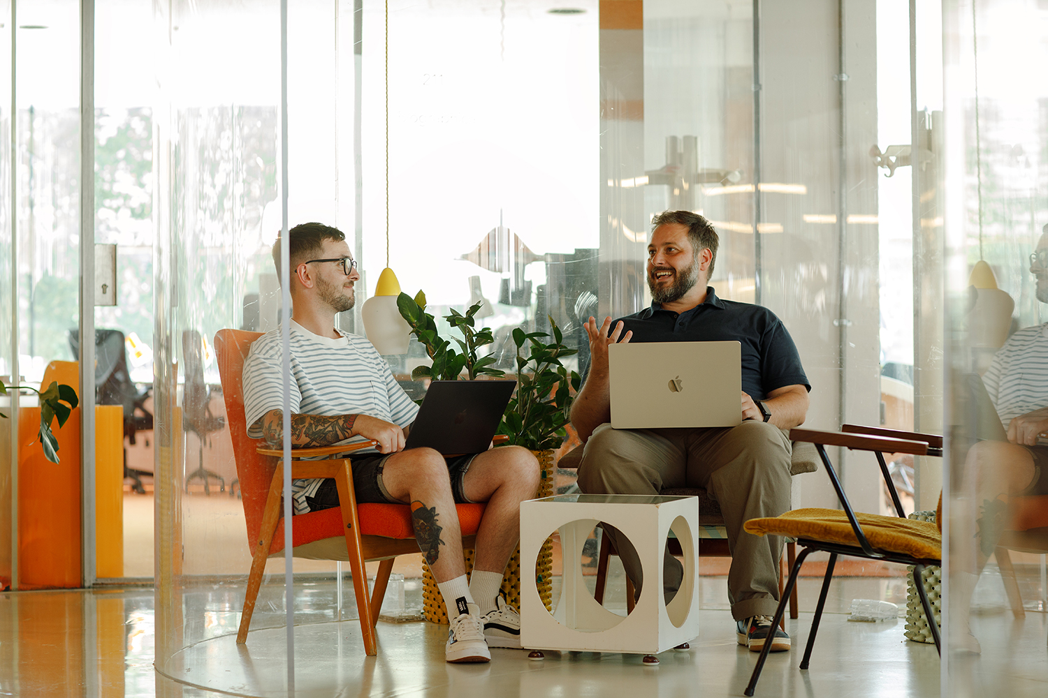Two people sitting in a modern office lounge area, chatting and smiling while working on laptops, with plants and colourful furniture around them.
