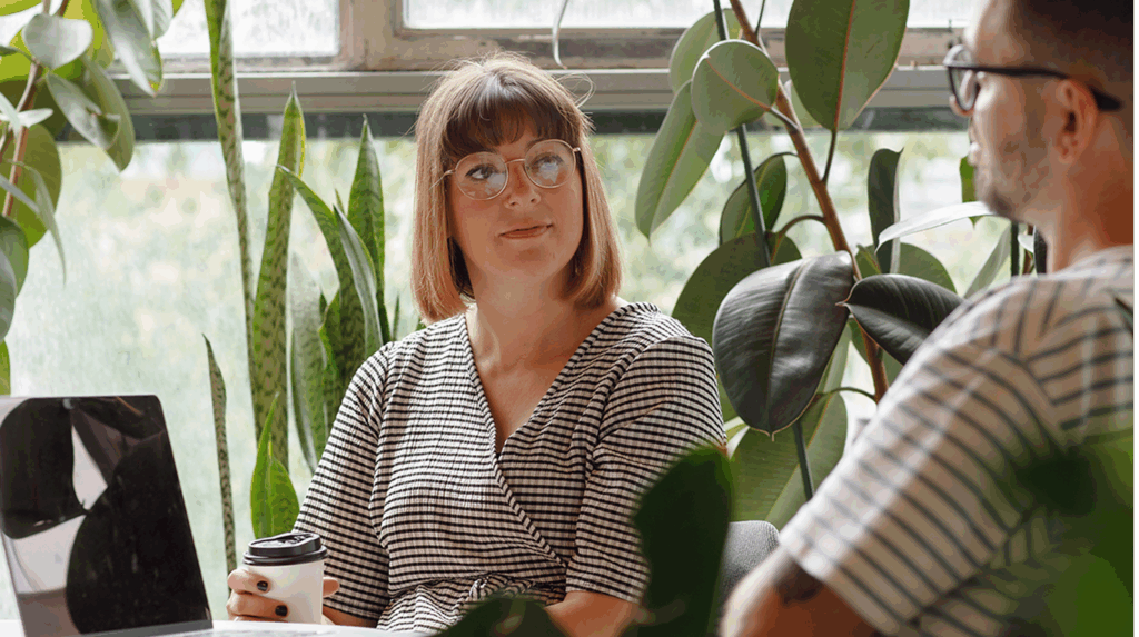 Two people in a Secure Innovation review at a desk, surrounded by large leafy plants, with one holding a takeaway coffee cup and a laptop in view.